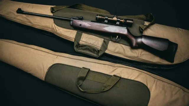 Bolt-action rifle with scope resting on a padded tan gun case with green straps, ready for transport.
